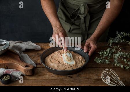 Hoher Winkel von anonymen Mann in Schürze mit Silikon-Spachtel zu Teig in Zinn zu verbreiten, während die Zubereitung Kürbiskuchen auf Holztisch Stockfoto