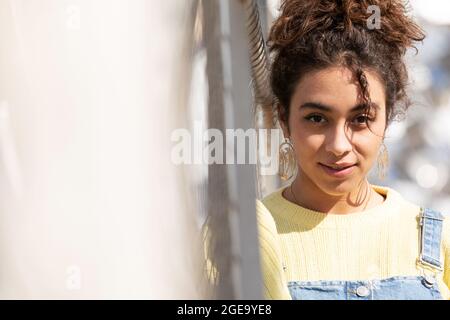 Selbstbewusste hispanische Teenager mit lockigen Haaren in Denim-Overalls und gelbem Sweatshirt mit Ohrringen, die auf die Kamera blicken, während sie sich auf raili lehnt Stockfoto