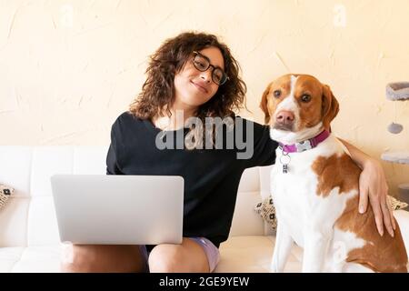 Junge hispanische Hündin streichelt Hund und Surfen im Internet auf Laptop, während Freizeit zusammen im Wohnzimmer verbringen Stockfoto