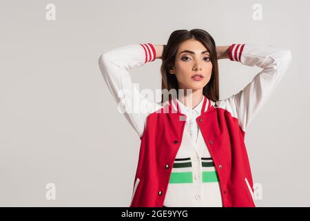 Brünette Frau in schwarz und weiß Jacke posiert mit Händen hinter dem Kopf isoliert auf grau Stockfoto