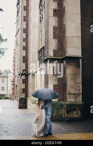 Stilvolle Braut und Bräutigam küssen unter Regenschirm auf dem Hintergrund der alten Kirche im Regen. Hochzeit in der Provence. Schöne Hochzeit Paar umarmt unter schwarz U Stockfoto