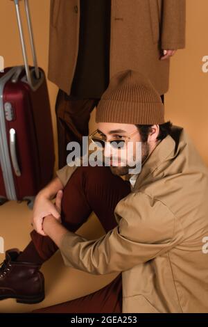 Sie Mann in Beanie und stilvolle Sonnenbrille sitzt in der Nähe verschwommene Frau mit Koffer auf beige Stockfoto