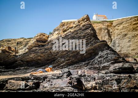 Zambujeira do Mar, Portugal - 28. Juni 2021: Ältere Frau, die am Strand auf den Felsen liegt und das Buch liest Stockfoto