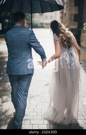 Stilvolle Braut und Bräutigam, die unter dem Regenschirm spazieren und die Hände auf dem Hintergrund der alten Kirche im Regen halten. Hochzeit in der Provence. Schönes Brautpaar walki Stockfoto
