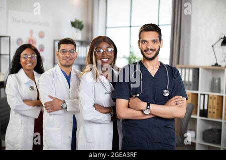 Porträt von vier selbstbewussten, multiethnischen Ärzten mit gekreuzten Armen, die in einem modernen Krankenhauszimmer in Reihe stehen. Multirassisches medizinisches Team, Gesundheitskonzept Stockfoto