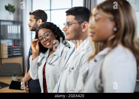 Profil im Hochformat. Multirassische Gruppe von vier Ärzten in Laborkittel, die in einer Reihe stehen. Konzentrieren Sie sich auf die hübsche afroamerikanische Dame in Brillen, lächelnd und in die Kamera schauend Stockfoto