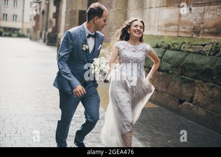 Schönes emotionales Hochzeitspaar, das lächelt und sich im Regen in der europäischen Stadt amüsieren kann. Hochzeit in der Provence. Stilvolle glückliche Braut und Bräutigam laufen auf dem Rücken Stockfoto