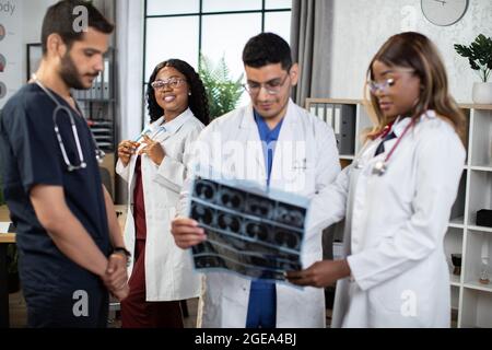 Multirassisches Ärzteteam, das den Röntgen-Scan eines Patienten diskutiert und im modernen Krankenhauszimmer gruppiert steht. Konzentrieren Sie sich auf lächelnde, hübsche afrikanische Frau, die dahinter steht Stockfoto