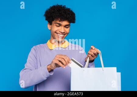 Fröhlicher afroamerikanischer Mann, der die Kreditkarte in eine Einkaufstasche auf blauem Himmel legt Stockfoto