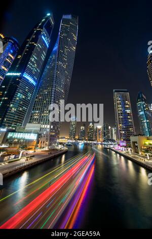 Der bunte Lichtpfad einer arabischen Dhow schlängelt sich durch den Yachthafen und unter den imposanten Gebäuden in Dubai. Stockfoto