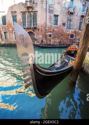 Das elegante Prow einer Gondel bocht im Wasser, als ein Gondolier im Hintergrund seine Passagiere in einem venezianischen Kanal entlang führt. Stockfoto