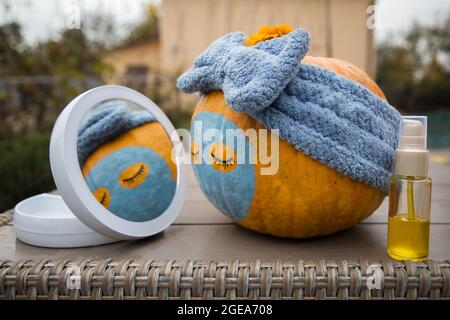 Großer orangefarbener Kürbis mit bemalten Augen, Lippen und einer Tonmaske vor einem runden Spiegel während einer Schönheitsbehandlung. Humor Schönheit Konzept. Selektiver FOC Stockfoto