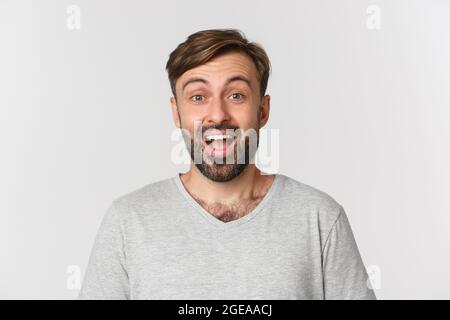 Nahaufnahme eines hübschen erwachsenen Mannes mit Bart, in grauem T-Shirt, lächelnd überrascht, auf weißem Hintergrund stehend Stockfoto