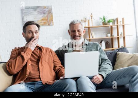 Lächelnder homosexueller Mann mit Laptop in der Nähe erstaunt Partner Stockfoto