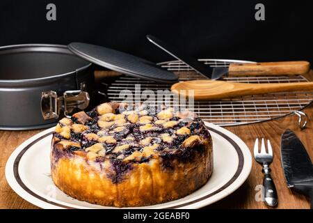 Seitenansicht von köstlichem hausgemachtem Heidelbeer und bröckelndem Käsekuchen in brauner Keramikplatte mit Holzlöffel, Servierschaufel auf Kühlregal, Metallgabel Stockfoto