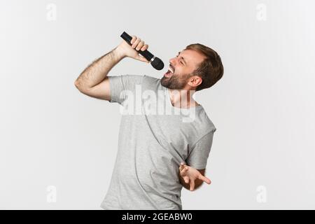 Schöner sorgloser Kerl, der Lied in Karaoke singt, Mikrofon hält und auf weißem Hintergrund steht Stockfoto