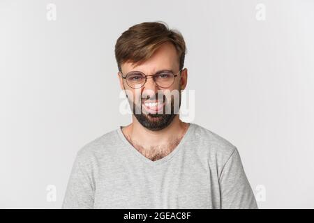 Nahaufnahme eines skeptischen bärtigen Mannes in Brille und grauem T-Shirt, grimazing angewidert und zeigt Zunge, stehend über weißem Hintergrund Stockfoto