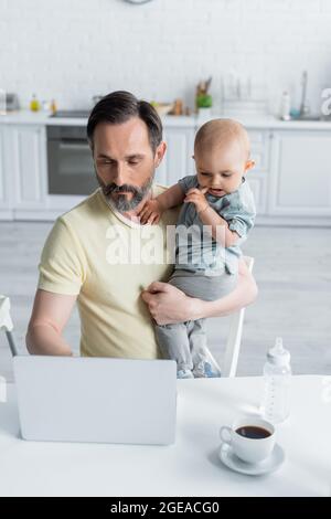 Reifer Vater hält Baby-Tochter und mit Laptop in der Nähe von Kaffee in der Küche Stockfoto
