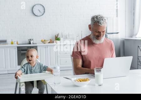 Baby Mädchen sitzt auf einem Hochstuhl in der Nähe lächelnder Vater mit Laptop Stockfoto
