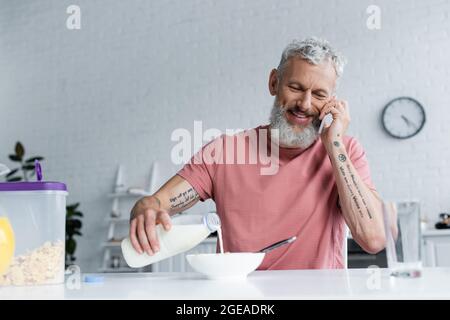 Reifer Mann gießt Milch in Maisflocken und spricht auf dem Smartphone Stockfoto