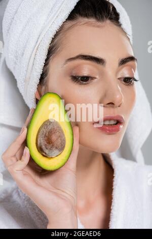 Nahaufnahme einer jungen Frau im Bademantel, die eine Hälfte der Avocado isoliert auf Grau hält Stockfoto