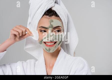Glückliche junge Frau im Bademantel entfernen Tonmaske mit Wattepad isoliert auf grau Stockfoto