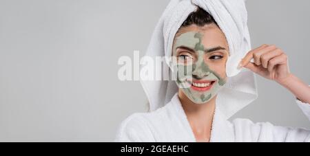 Glückliche junge Frau im Bademantel entfernen Tonmaske mit Wattepad isoliert auf grau, Banner Stockfoto