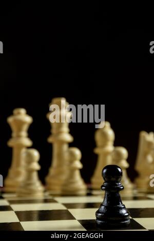 Auf einem Schachbrett im Vordergrund ist ein schwarzes Schachstück Pawn. Hinter ihr sind eine Reihe von weißen Formen verschwommen. Hinter den Figuren ist ein schwarzer Hintergrund. Stockfoto