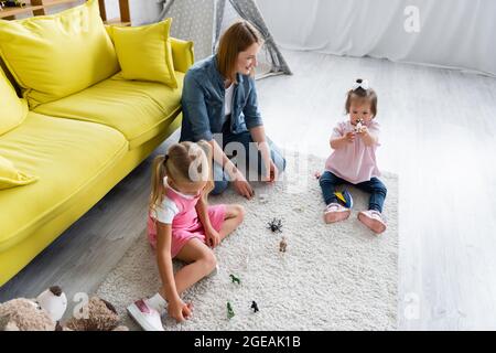 Hochwinkelansicht der Kindergärtnerin, die ein Kleinkind mit Down-Syndrom betrachtet, das mit Spielzeug spielt, in der Nähe eines verschwommenen Vorschulkindes im Spielzimmer Stockfoto