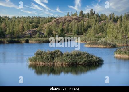 Ein See mit einer Ebenen Oberfläche und Reflexionen gegen den blauen Himmel und weiße Wolken. Entstanden aus einem alten Steinbruch überflutet, Gruppen von Inseln überwuchert Stockfoto