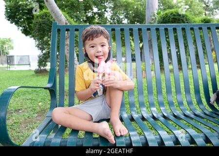 Netter kleiner Junge, der auf einer Parkbank sitzt und Eis isst Stockfoto