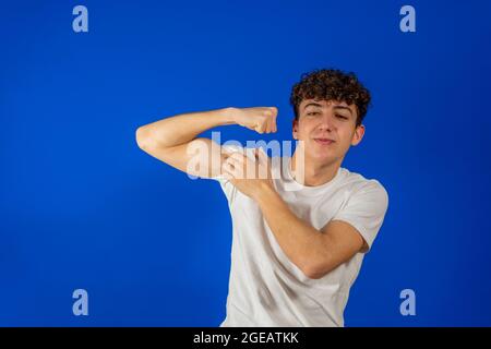 Stolzer Mann, der auf seinen Bizeps zeigt und auf die Kamera schaut. Porträt eines hübschen jungen Hipster-Lockenmannes in klassischem, lässigem weißen Hemd. Innenaufnahme im Studio Stockfoto