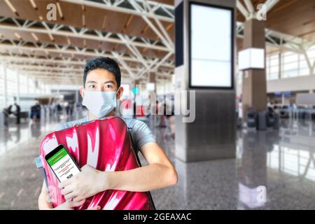 Glücklicher Reisender mit digitalem Impfpass am Telefon und Umarmung seines Gepäcks am Flughafen für seine erste Reise nach den COVID-19-Beschränkungen Stockfoto