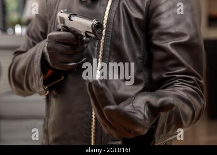 Bewaffneter Raub, Schütze mit einer Pistole in der Hand mit Handschuhen. Mann in schwarzer Lederjacke, der mit einem Gewehr anvisieren wollte, um Geld bat, Nahaufnahme. Stockfoto