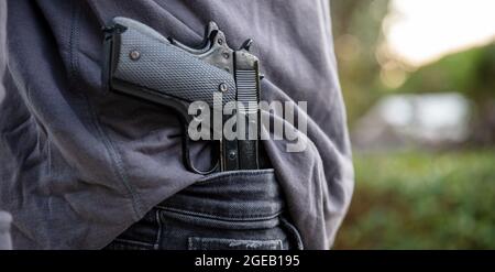Bewaffneter Mann, der eine Pistole in seinem Jeans-Bund trägt, verwischen den Hintergrund der Natur im Freien. Konzept von Bedrohung, Gewalt und Gefahr Stockfoto