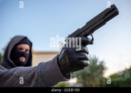 Mann, der mit einer Pistole anvisierte, Natur im Hintergrund, Nahaufnahme. Killer mit Sturmhaube, hält eine Waffe in der Hand mit Handschuhen, bewaffnete Räuber Konzept Stockfoto
