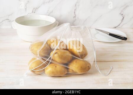 Rohe Bio-Kartoffeln in einem wiederverwendbaren Öko-Netzbeutel auf einem weißen Holztisch. Recyceltes Nylon produziert Taschen für den Einkauf und die Lagerung von Lebensmitteln. Keine Verschwendung. Stockfoto