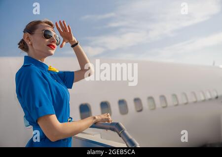 Porträt einer eleganten Luftverkehrsleiterin in blauer Uniform und einer Sonnenbrille, die die Hand am Kopf hält und weit weg schaut und tagsüber auf der Lufttreppe steht Stockfoto