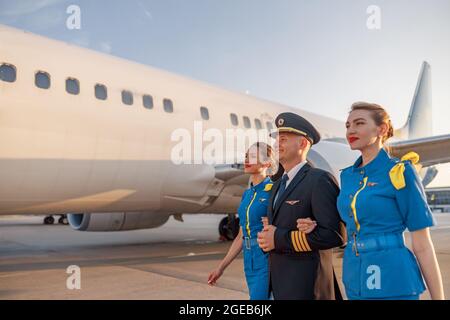 Aufgeregter männlicher Pilot, der bei Sonnenuntergang mit zwei Flugbegleitern in blauer Uniform vor einem Flugzeug im Terminal läuft Stockfoto