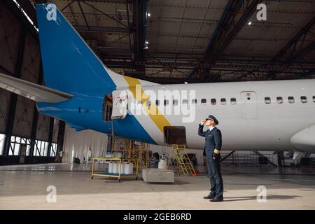 Stolzer Pilot in Uniform schaut weg, passt seinen Hut an und steht vor einem großen Passagierflugzeug im Flughafenhangar Stockfoto