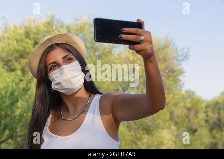 Junge attraktive weibliche Reisende im Hut macht Selfie vor Hintergrund, trägt schützende medizinische Maske, um sich vor Coronavirus zu schützen. Pandemie c Stockfoto