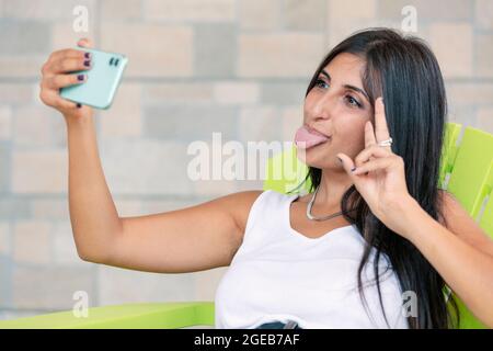 Junge Frau macht dummes Gesicht und zeigt Zunge auf Smartphone, während Selfie zu machen. Glückliche Person, die Spaß hat und V-Zeichen mit der Hand macht. Mädchen nimmt f Stockfoto