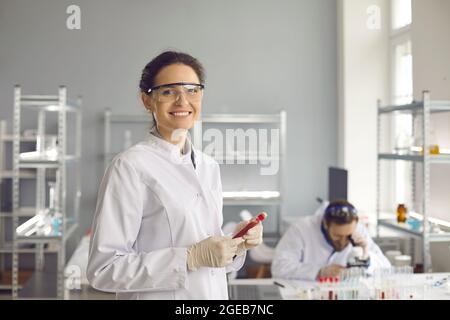 Porträt eines glücklichen, lächelnden Wissenschaftlers in Laborkittel und Brille mit dem Bluttestrohr Stockfoto