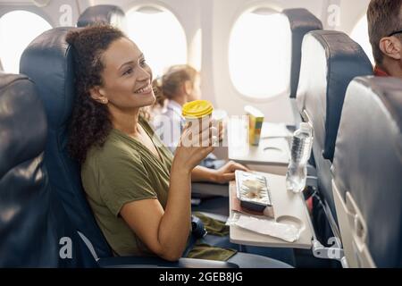 Glückliche weibliche Fahrgastin trinkt Kaffee und lächelt, während eine Flugbegleiterin das Mittagessen an Bord serviert Stockfoto