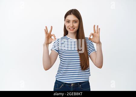 Lächelndes Mädchen zeigt okay Zeichen in Zustimmung, steht über weißem Hintergrund in lässiger Kleidung Stockfoto