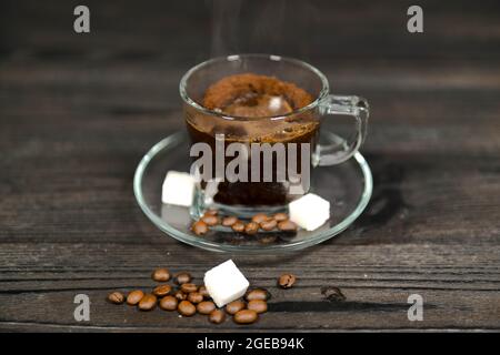 Eine Tasse aus Glas und eine Untertasse auf einem dunklen Holzhintergrund. Es ist mit einer braunen Flüssigkeit gefüllt, es ist gebrühter Kaffee. Es gibt Zuckerwürfel und Kaffee Bea Stockfoto