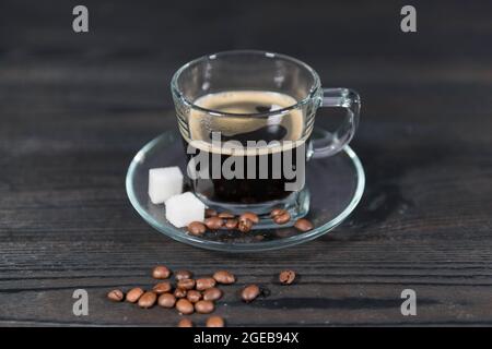 Eine Tasse aus Glas und eine Untertasse auf einem dunklen Holzhintergrund. Es ist mit einer braunen Flüssigkeit gefüllt, es ist gebrühter Kaffee. Es gibt Zuckerwürfel und Kaffee Bea Stockfoto