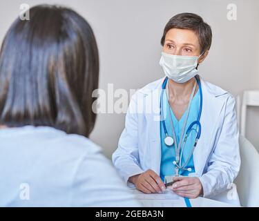 Professionelle reife Frau Arzt in Gesichtsmaske Beratung weibliche Patientin während covid-19 Ausbruch Stockfoto