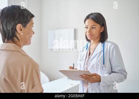 Lächelnde afrikanische Ärztin mit Klemmbrett in den Händen Gefühl der Krankengeschichte von reifen weiblichen Patienten Stockfoto