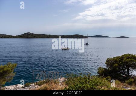Segelboote, die sich in der Nähe der Küste von Rogoznica an der Adria vorbeiziehen, folgen der grünen Küste, die von dichtem Pinienwald bedeckt ist Stockfoto
